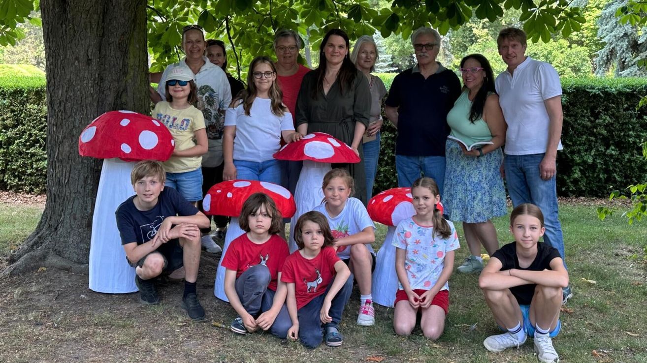 A group of people, including children and adults, stand and crouch under a tree. Some wear mushroom hats. One woman holds a book. They seem to be posing for a photo.