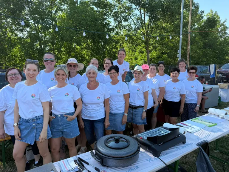 A group of women wearing white shirts with logos are posing for a photo. They are standing in front of a table with a pot, paper roll, and knives. Behind them are trees, lights, and a grassy area.