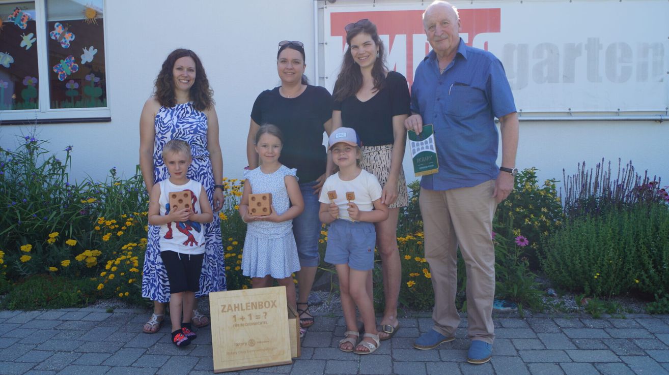 A family and their children pose for a picture in front of a building with flowers, holding wooden boxes with numbers.