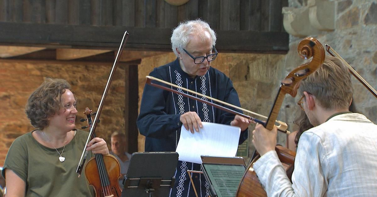 A man holding a violin bow looks at a piece of paper. Violinists in the background are playing while another person holds a cello.