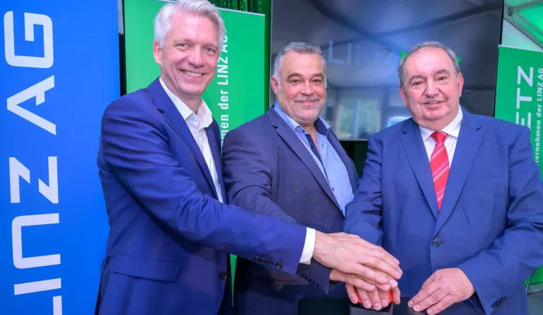 Three men in business suits are smiling and shaking hands in front of a green sign with white letters.