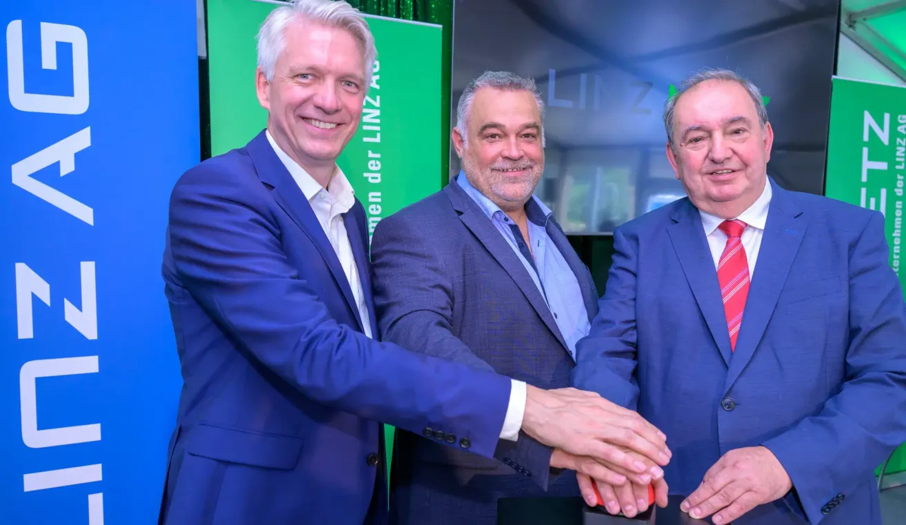 Three men in business suits are smiling and shaking hands in front of a green sign with white letters.