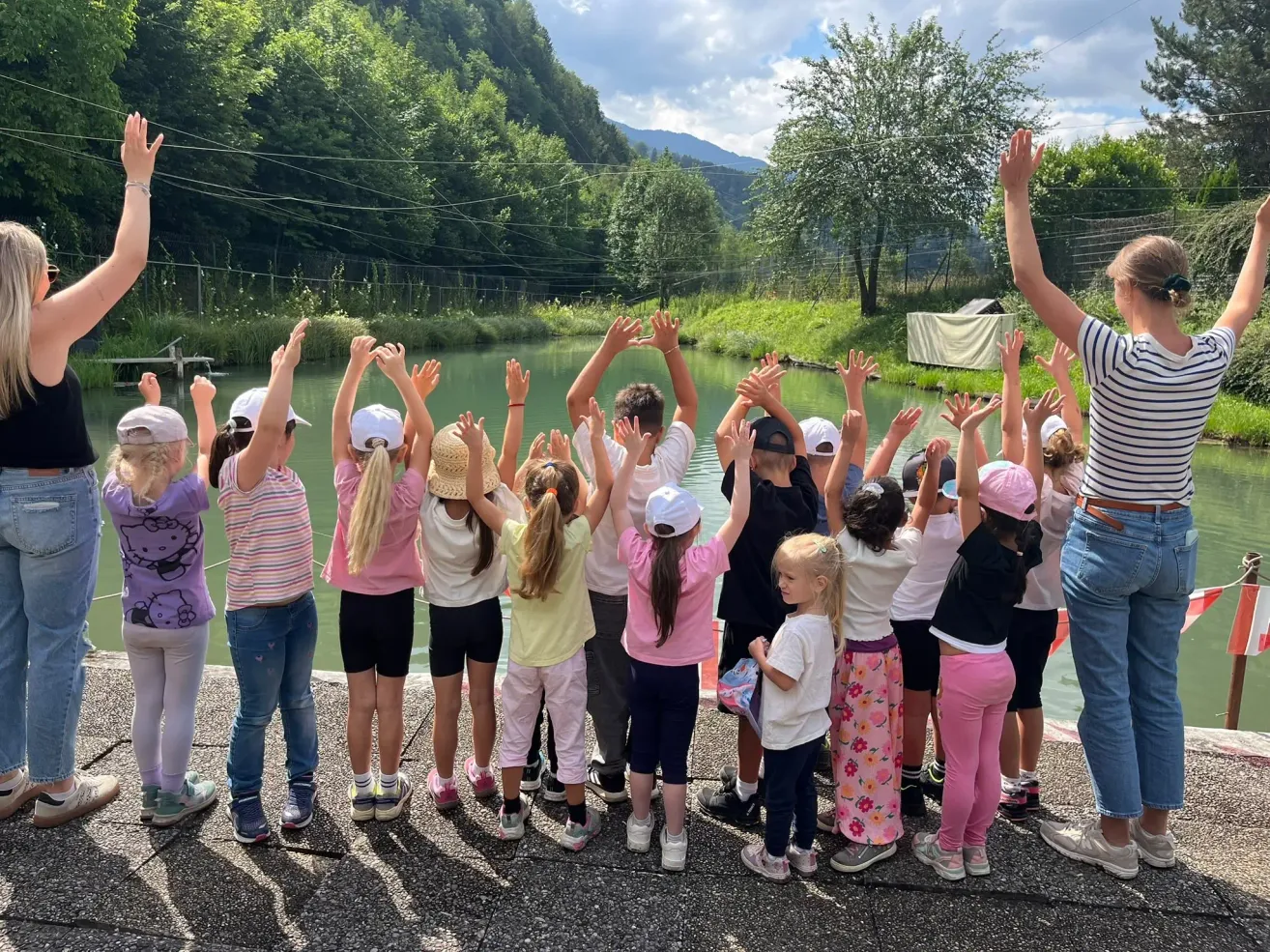 A group of children stand by the water with raised hands, likely posing for a photo. They wear various hats and shoes. Behind them is a grassy area with trees and a mountain landscape.
