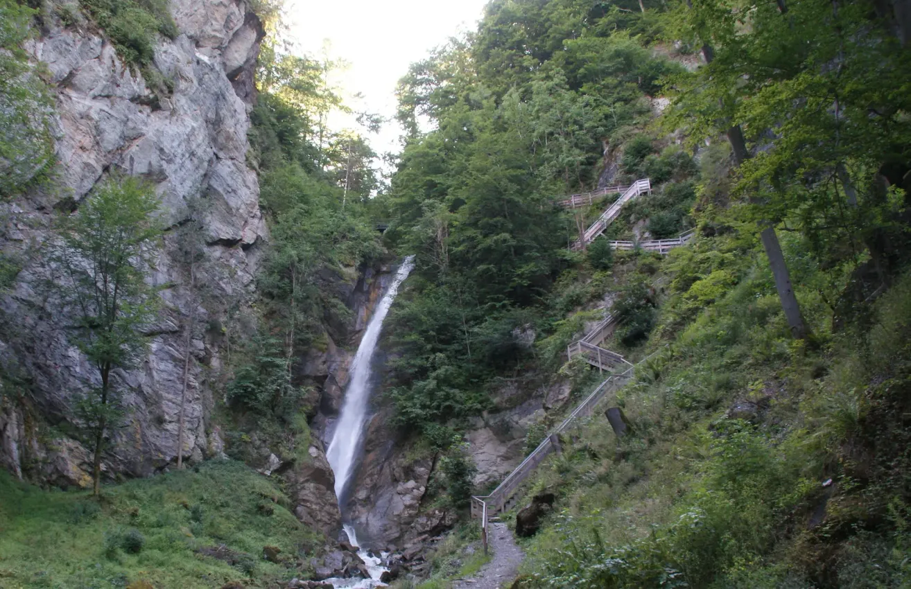 A waterfall cascades down a rocky cliff, surrounded by lush greenery. Wooden staircases lead up the steep terrain. The water flows into a stream at the base.