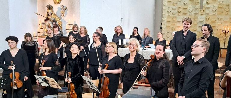 A group of musicians in black clothing stand in a church, holding various instruments, with candles and a statue in the background.