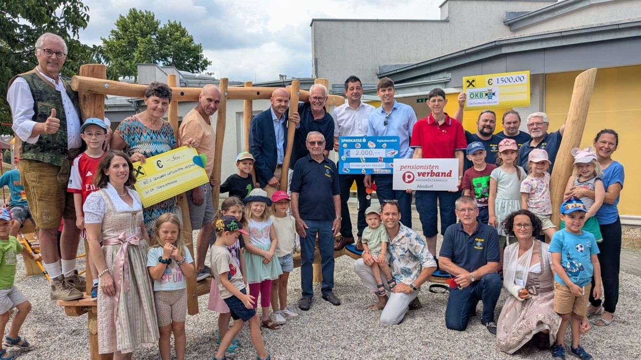 A group of adults and children pose for a photo in front of a playground. The adults hold a check for 2,000 euros and a poster reading 'Pensionisten Verband Absdorf'.