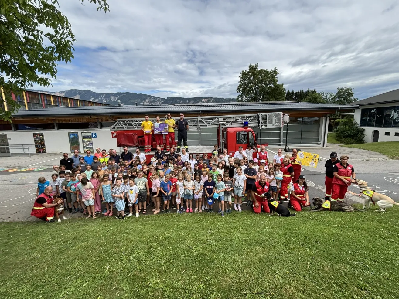 A large group of people, including children, adults, and firefighters, gather around a red fire truck in front of a building with mountains in the background.