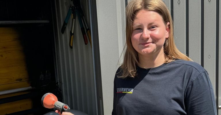 A woman with blonde hair wearing a black shirt stands in a shed, holding a tool.