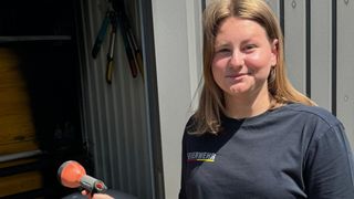 A woman with blonde hair wearing a black shirt stands in a shed, holding a tool.