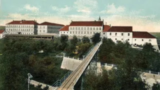 An old photograph shows a large, historical building with red roofs and multiple windows, a bridge in front, and a lush green landscape.