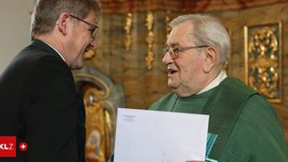 Two men are conversing inside a church. An older man holds a piece of paper, while the younger man wears glasses and a suit.