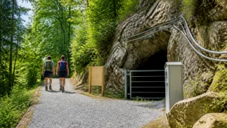 Two hikers, wearing backpacks, walk on a gravel path beside a rocky cliff, with a tunnel entrance nearby.