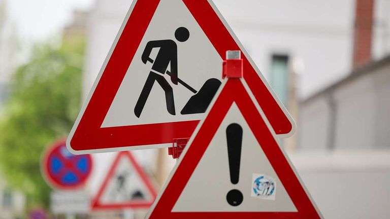 A red and white triangle warning sign depicts a person digging with a shovel. Below it is another triangle sign with a black exclamation mark.
