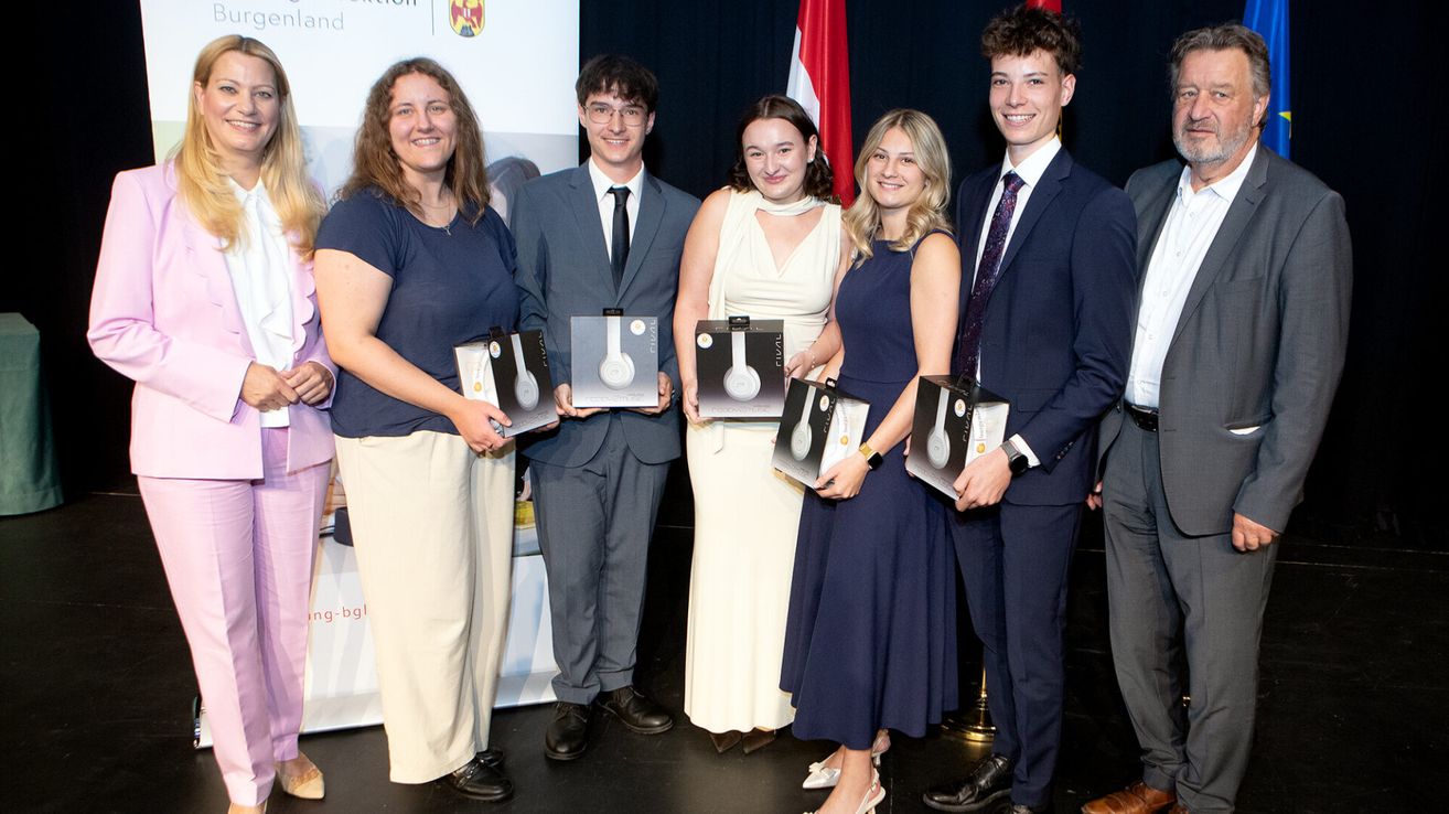 Five young adults stand on a stage, holding boxes. Two women and three men pose for a photo. They are smiling and appear to be celebrating an award. Behind them, a banner with text and a flag are visible.