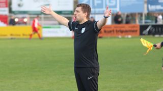 A soccer referee with arms outstretched on a field. He wears a black uniform with a Nike logo on the shorts. Behind him is a blurred background with people and a banner.