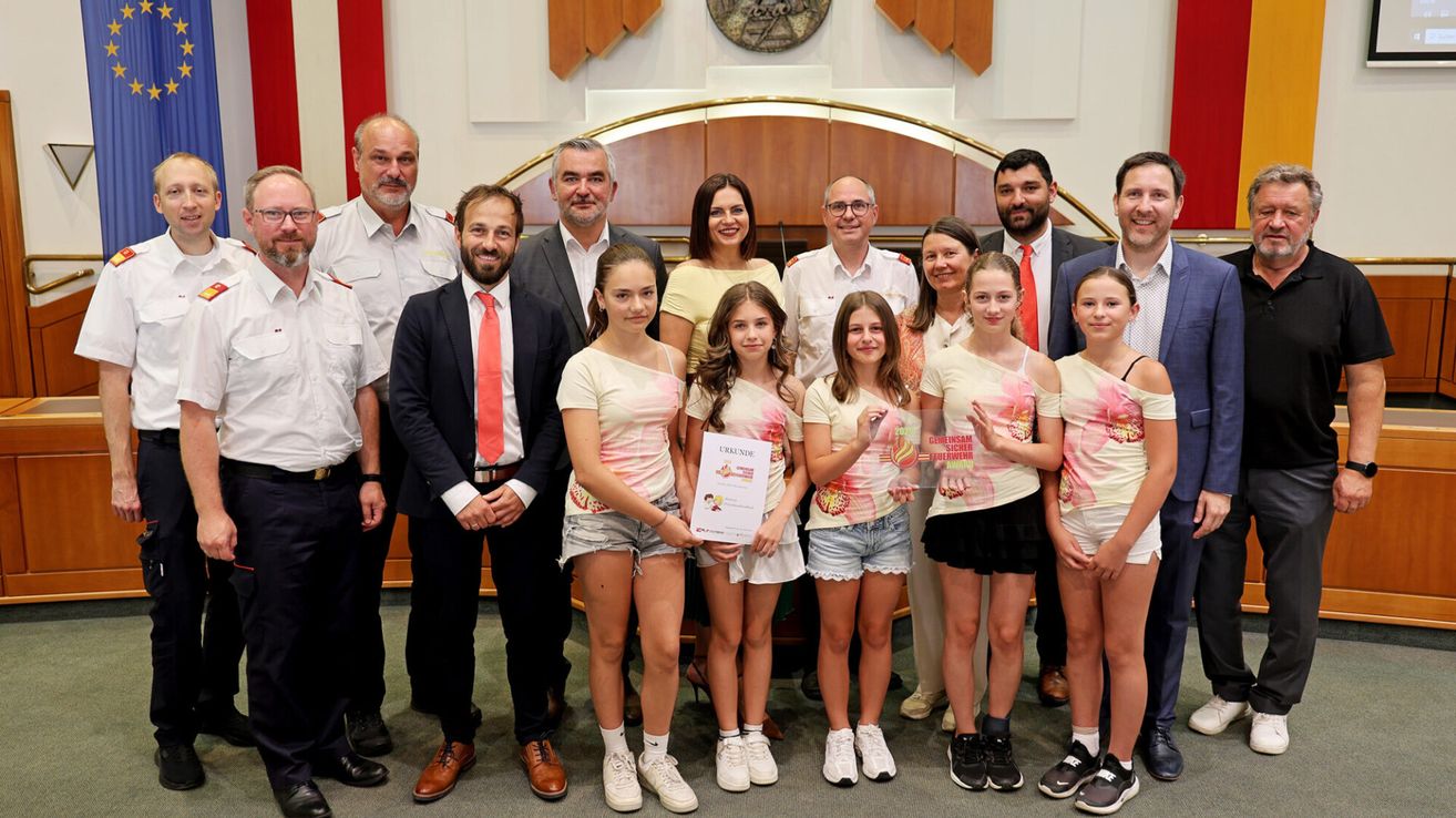 Group photo with several adults and young girls, holding a certificate, posing for a picture in front of a wooden structure.
