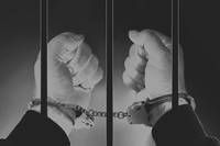 Two hands, one clenched into a fist and the other open, are held behind bars. Both wrists are restrained by handcuffs.