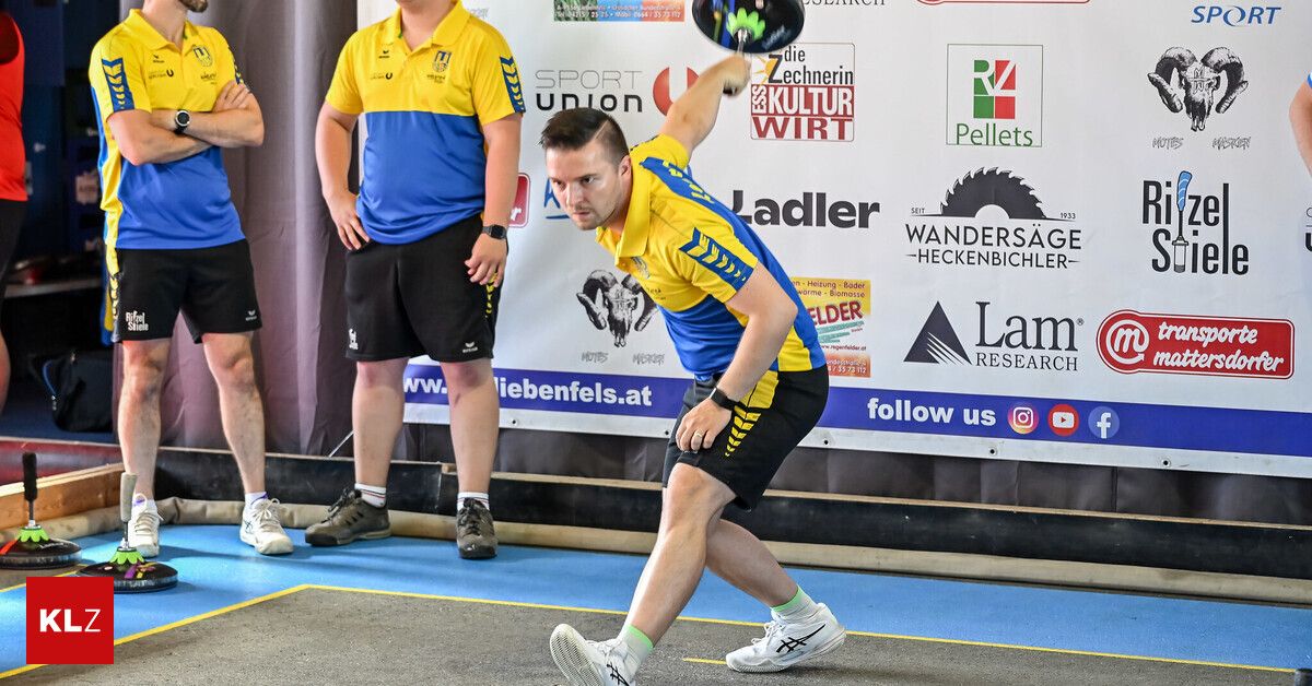 A man in a yellow and blue shirt throws a disc in a disc golf competition. A man in a yellow and black shirt watches in the background.