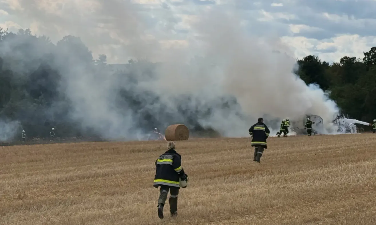 Feuerwehrleute auf einem Feld mit einem brennenden Heuballen und aufsteigendem Rauch im Hintergrund.