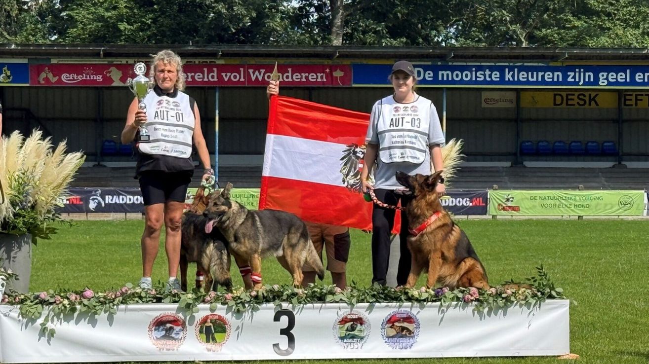 Zwei Frauen stehen auf einem Podest mit ihren Deutschen Schäferhunden, eine hält eine Trophäe und die andere eine Flagge. Sie sind von Blumen und einer Nummer 3 auf dem Boden umgeben.