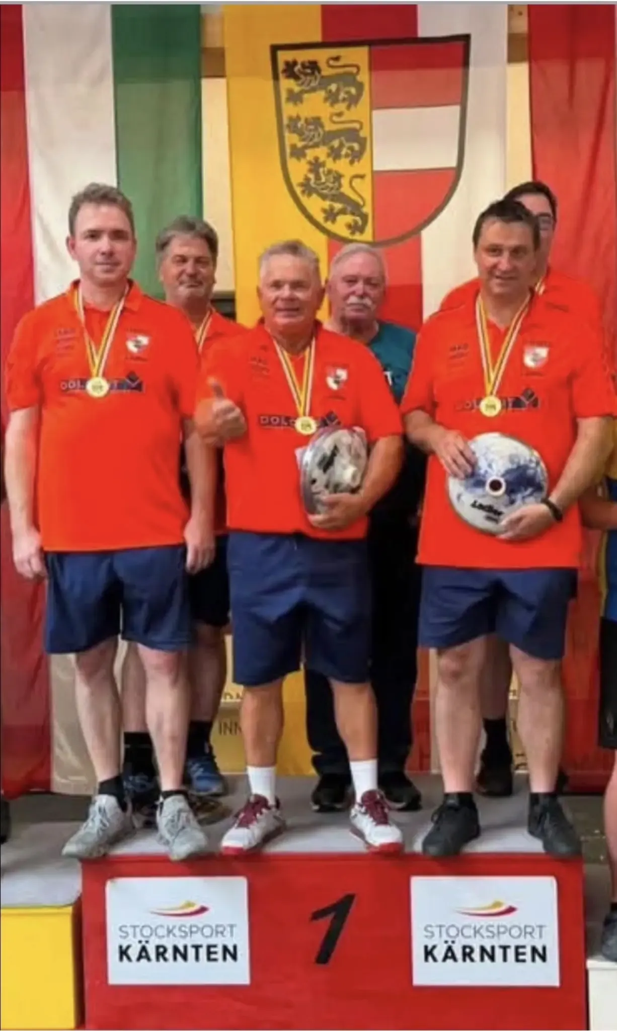 Five men in orange shirts and blue shorts stand together, holding medals and trophies. They are smiling and appear to be celebrating a victory. Behind them, a colorful flag with a logo is visible.