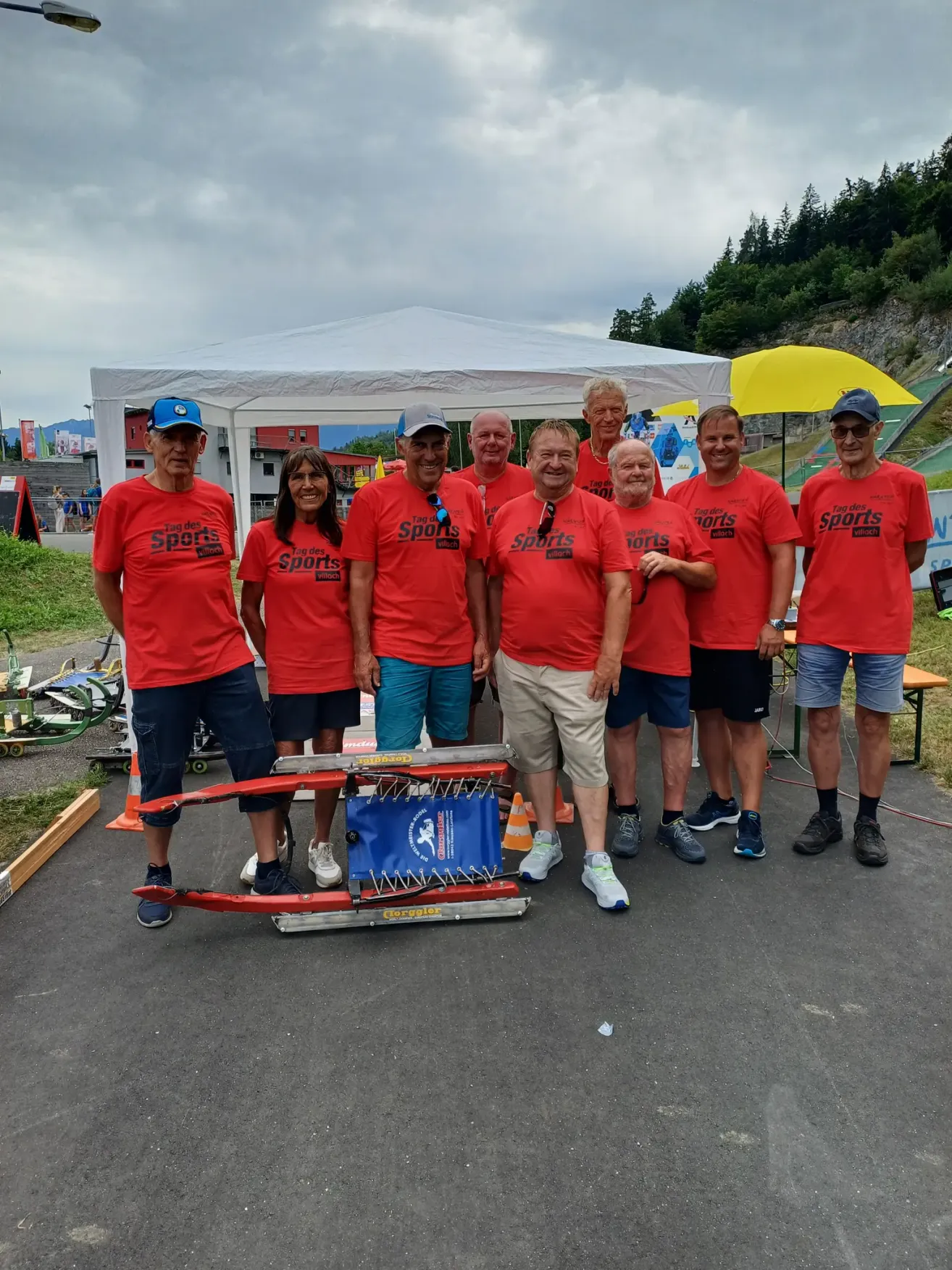 Eine Gruppe von Menschen in roten Sportshirts steht vor einem Zelt, mit einem roten Schlitten auf dem Boden und einem gelben Schirm dahinter.