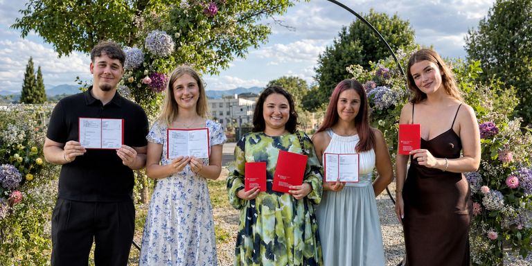 Five individuals, four women and one man, are standing in an outdoor area, holding red booklets, and smiling for a photograph. They are surrounded by trees and plants.