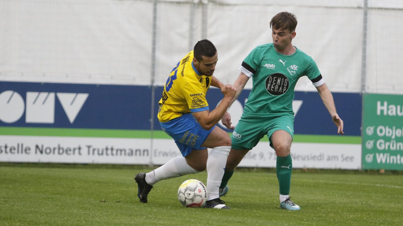 Zwei Fußballspieler sind in einem Spiel engagiert, einer in einem gelben und blauen Trikot und der andere in einem grünen Trikot mit einem Sponsorenlogo auf der Brust. Sie versuchen beide, den Ball auf dem Feld zu kontrollieren.