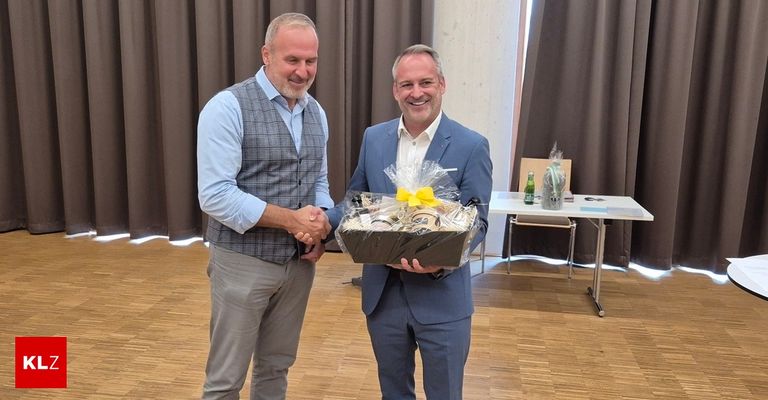 Two men in formal attire are shaking hands while one holds a gift basket with a yellow ribbon. The setting appears to be a professional event.