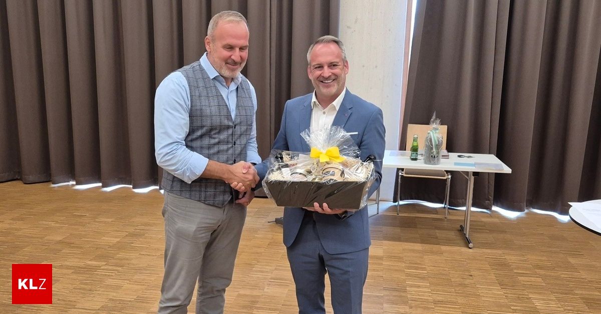 Zwei Männer in formeller Kleidung schütteln sich die Hände, während einer einen Geschenkkorb mit einem gelben Band hält. Der Ort scheint ein professionelles Ereignis zu sein.