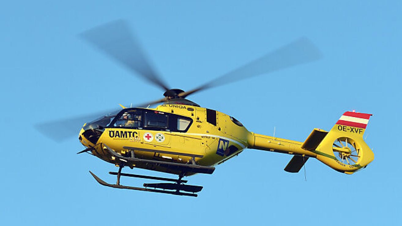 A yellow helicopter with the text 'OAMTC' and a red cross symbol flies in the sky. It has a black rotor and a blue tail.