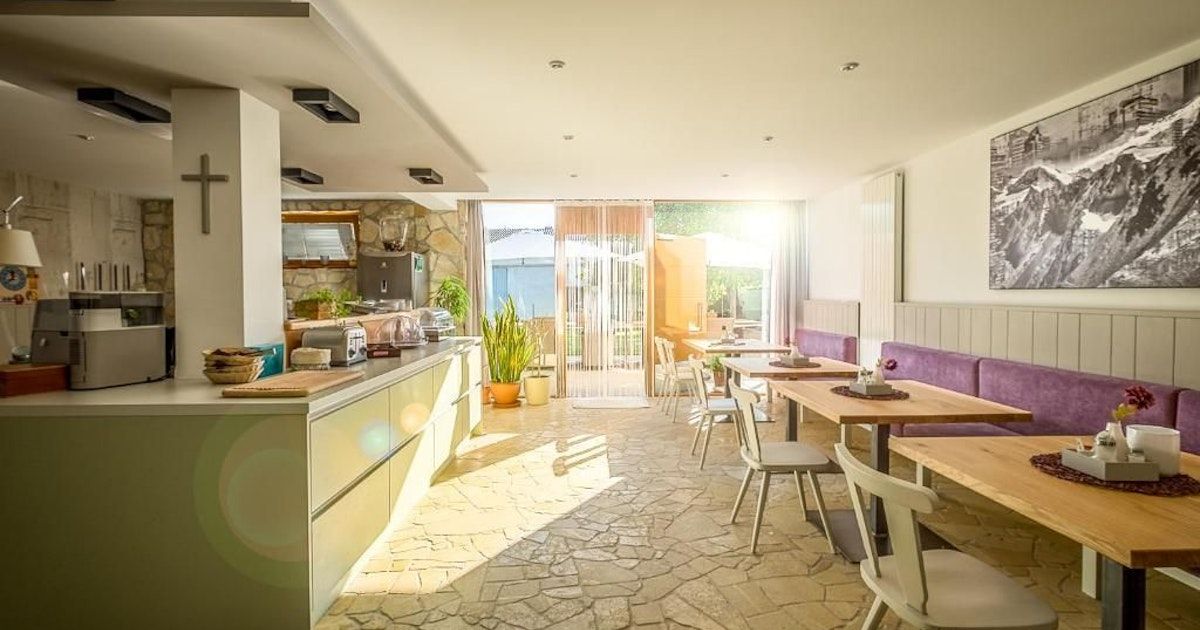 An interior view of a restaurant with a stone floor, wooden tables, and a kitchen area with a counter and appliances.
