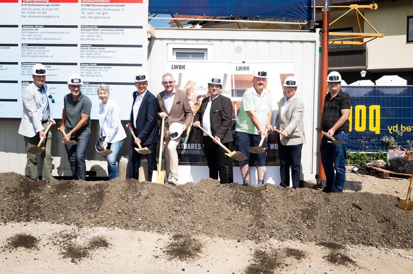 A group of people in hard hats and shovels are gathered for a groundbreaking ceremony in front of a building.