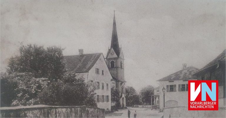 A black and white photograph of a church with a steeple and clock, situated in a small town with buildings and trees. Two people are walking on the street.