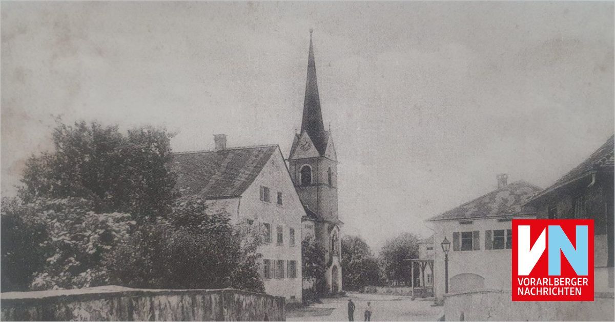A black and white photograph of a church with a steeple and clock, situated in a small town with buildings and trees. Two people are walking on the street.