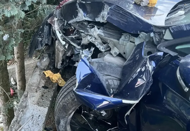 A severely damaged blue car with a crumpled front end and exposed engine components lies on the side of the road.