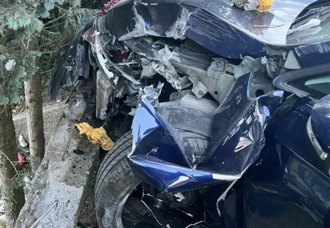 A severely damaged blue car with a crumpled front end and exposed engine components lies on the side of the road.