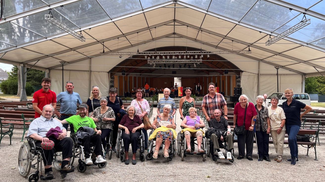 A group of elderly individuals, some in wheelchairs, pose for a photo in front of a stage under a tent.