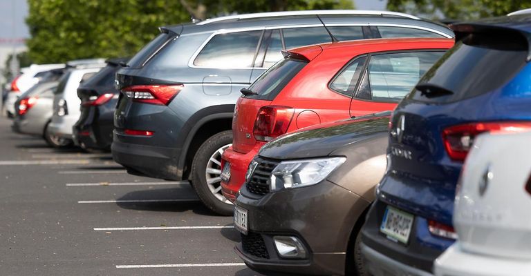 A parking lot with several cars parked in a row, including a gray SUV, a red hatchback, and a blue car. The cars are parked in a straight line with white markings on the ground.