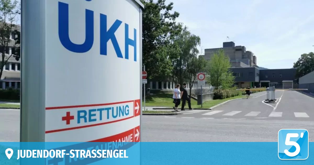 Ein großes Schild für UKH Rettungsdienst ist an der Straßenseite sichtbar. Zwei Personen gehen auf dem Bürgersteig vor einem Gebäude.