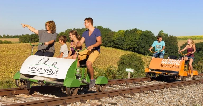 A group of people ride on a pedal-powered rail cart on a track with a scenic background.
