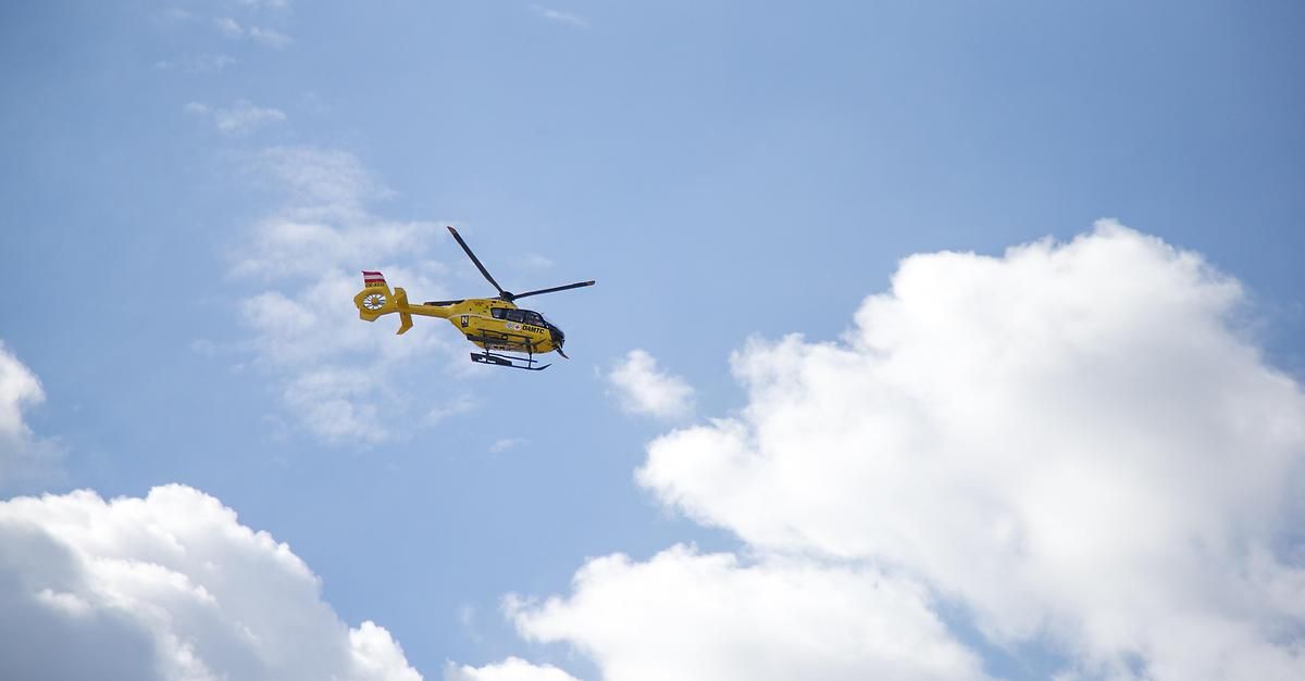 Ein gelber Hubschrauber mit rotem Kreuzsymbol fliegt am blauen Himmel mit weißen Wolken.