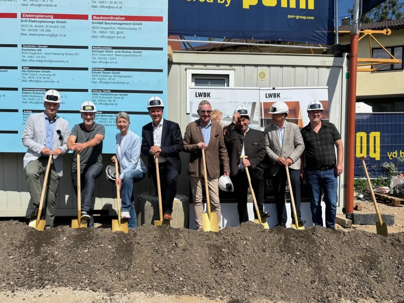 A group of people in hard hats and suits are standing in a dirt area, holding shovels, and posing for a photo. Behind them, a blue banner with text and logos is attached to a building.