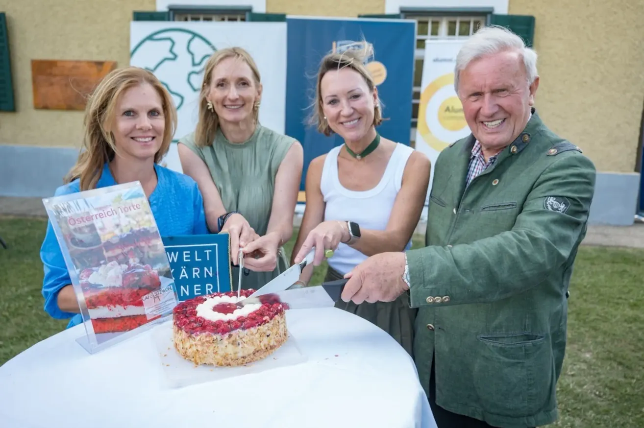 Vier Frauen und ein Mann lächeln, während sie einen Kuchen mit einem Messer auf einem Tisch schneiden. Der Kuchen hat eine rote Spitze und ein blaues Buch liegt auf dem Tisch.
