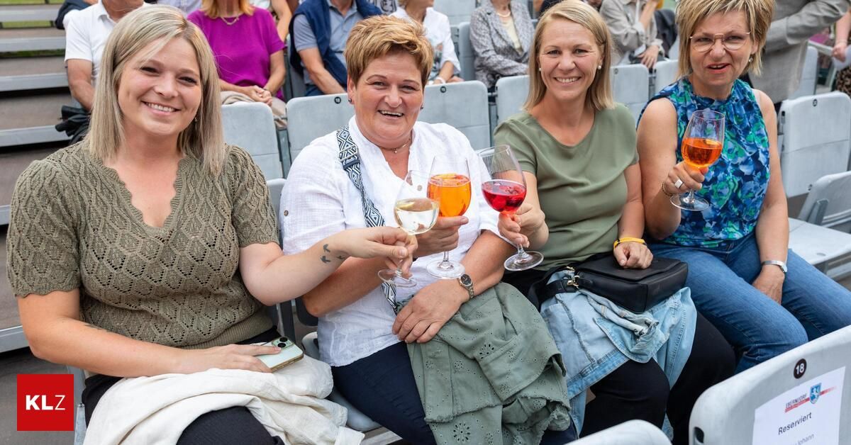 Four women sitting in a stadium, smiling and holding wine glasses. The woman in the middle is holding two glasses, one with a red drink and the other with a yellow drink. The woman on the right is holding a glass with a red drink. The woman on the left is holding a glass with a yellow drink.