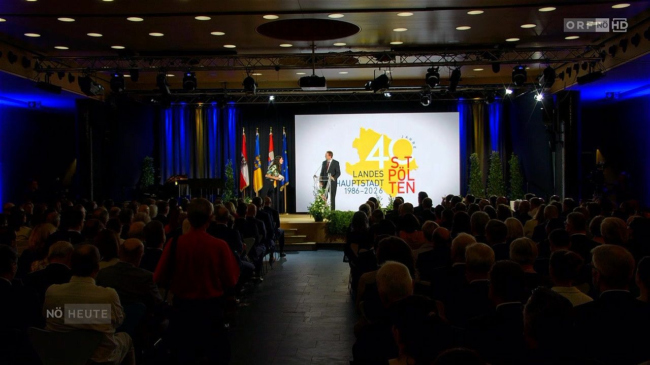 A formal event in a conference hall with a large screen displaying a yellow map and text. A man and a woman stand on stage with flags behind them.