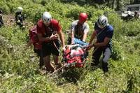 Four individuals in helmets and safety gear are carrying a stretcher through a dense, grassy area. The person on the stretcher appears to be injured.
