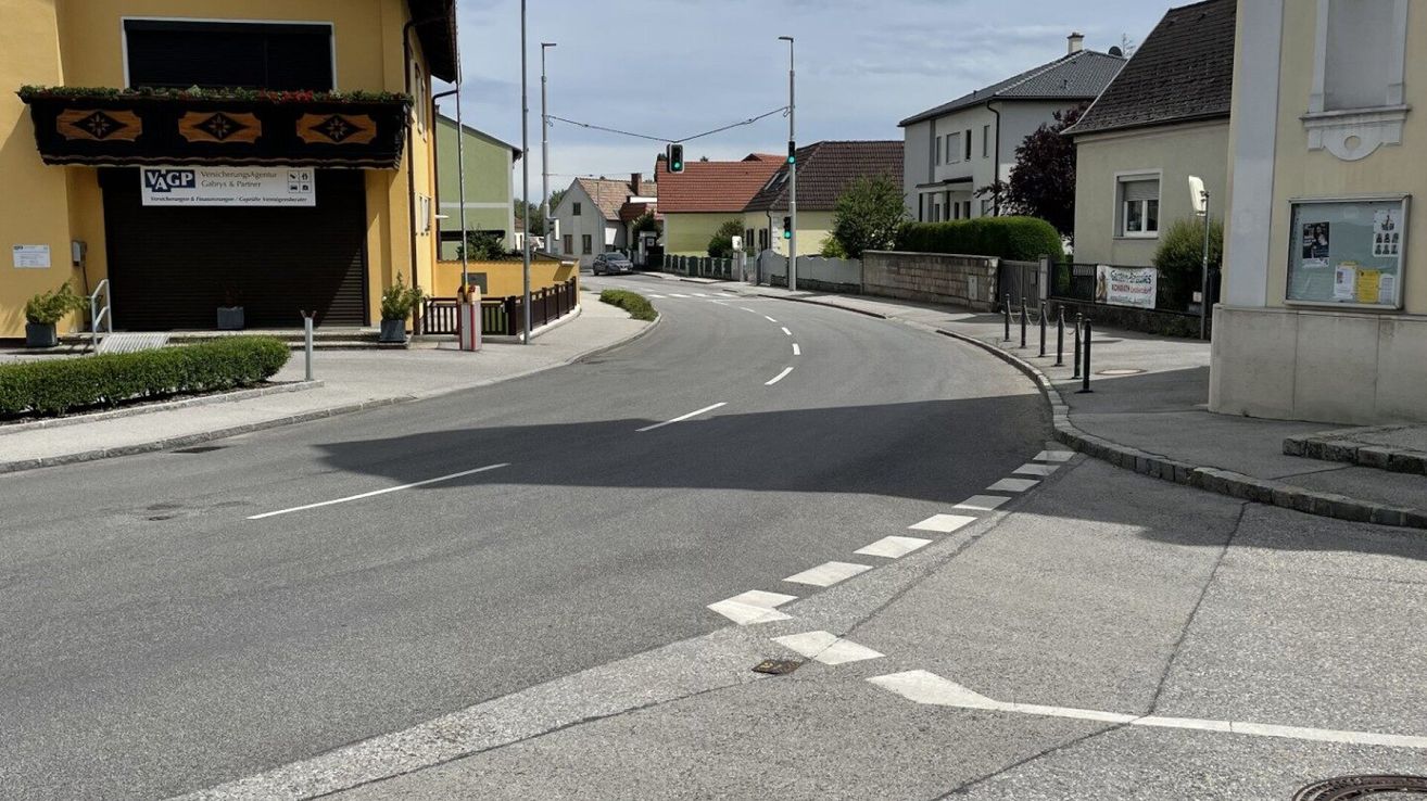 Eine leere Straße mit weißen Fahrbahnmarkierungen und einer Ampel. Gebäude in verschiedenen Farben und Stilen säumen die Straße.