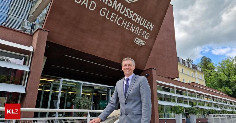 A man in a suit stands smiling in front of the Bad Gleichenberg building, a modern architectural structure with a brown facade and large text.