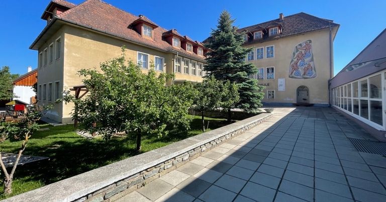 Ein gepflegter Innenhof eines Gebäudes mit einem gepflasterten Weg, einem kleinen Garten mit Bäumen und einer Wandmalerei an der Wand.